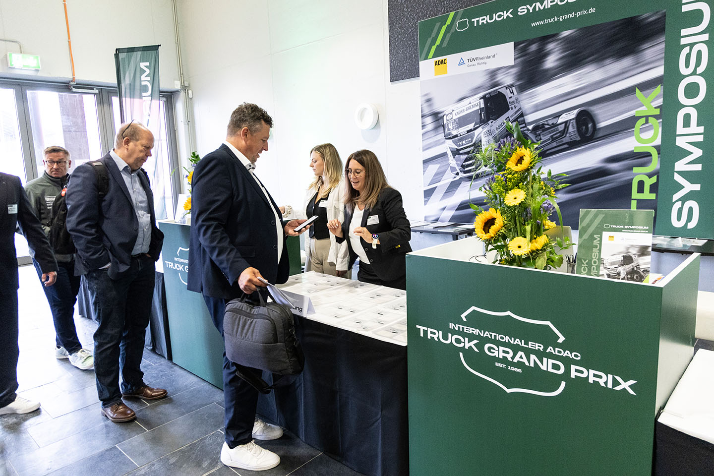 Eine Gruppe von Menschen steht an einem Empfangstisch beim Internationalen ADAC Truck Grand Prix. Zwei Frauen hinter dem Tisch begrüßen die Gäste. Der Tisch ist mit einem Blumenarrangement dekoriert.