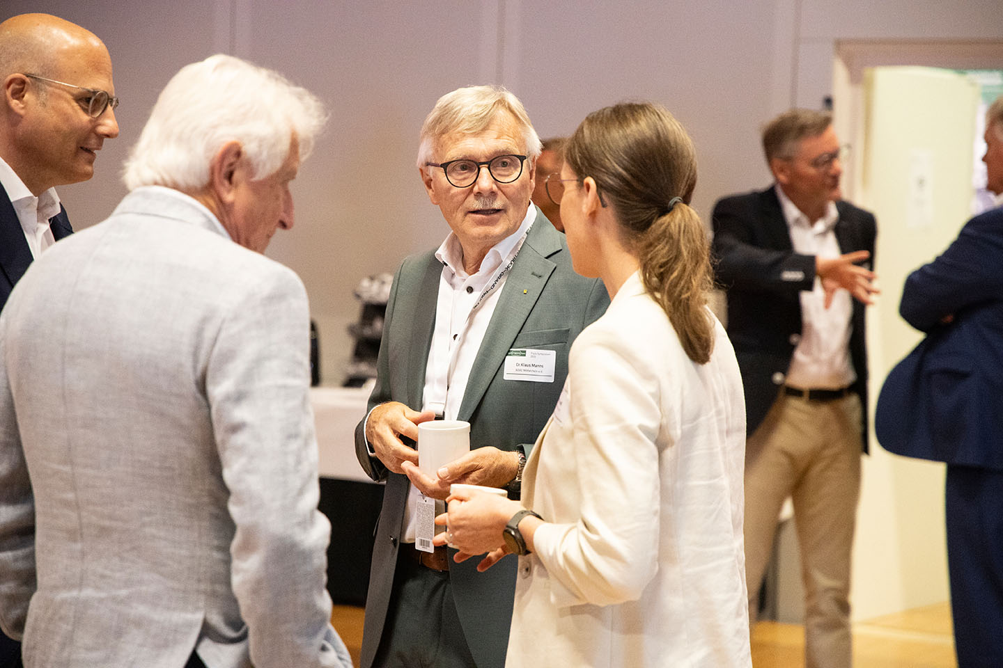 Eine kleine Gruppe von Personen steht im Gespräch miteinander. Eine Person hält eine Tasse in der Hand. Sie scheinen in einer Konferenzumgebung zu sein.