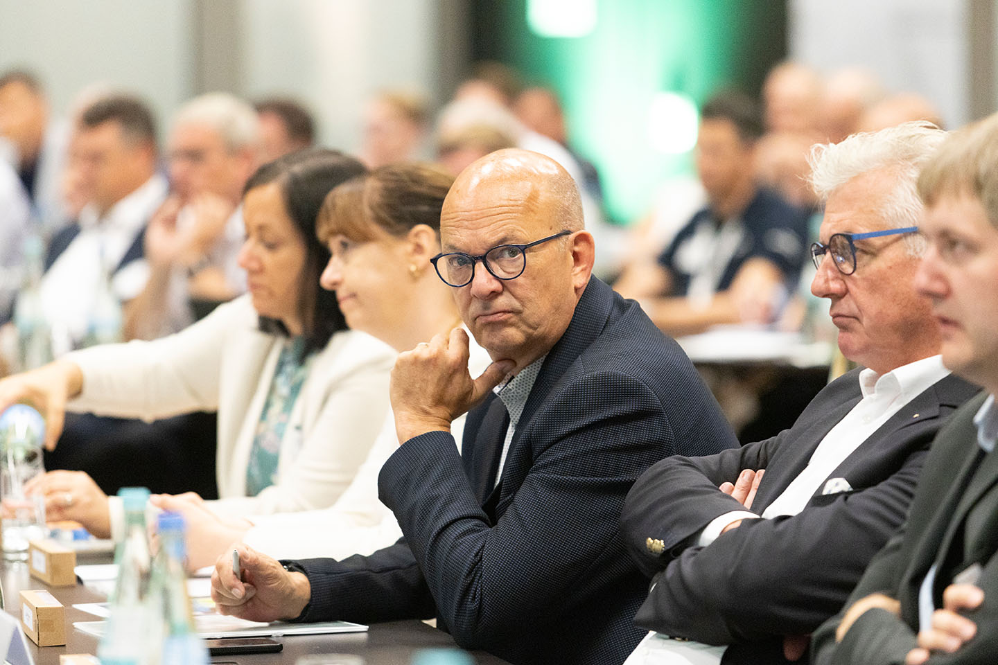 Mehrere Personen sitzen in einem Konferenzraum. Ein Mann im Vordergrund schaut aufmerksam nach vorne. Die Teilnehmer sind in Business-Kleidung.