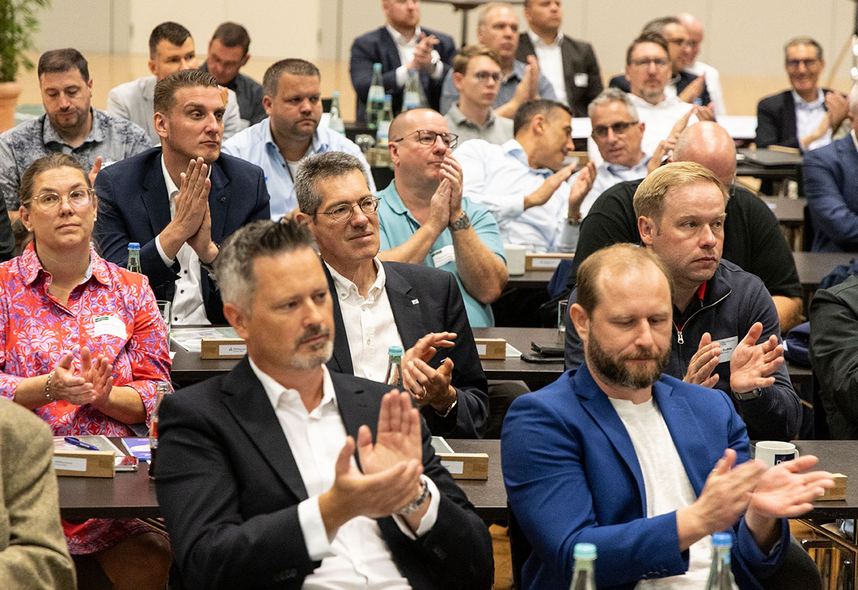 Ein Publikum in einem Konferenzraum applaudiert. Die Teilnehmer tragen Business-Kleidung und wirken interessiert an der Präsentation.