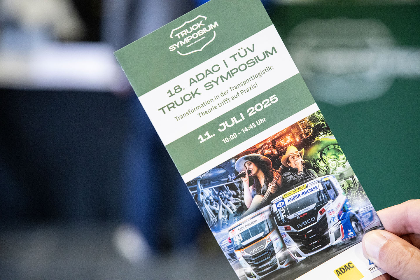 Eine Hand hält ein Programmheft des 18. ADAC | TÜV Truck Symposiums. Auf dem Heft sind das Datum, die Uhrzeit und Bilder von Lastwagen und Personen mit Cowbohüten abgebildet.