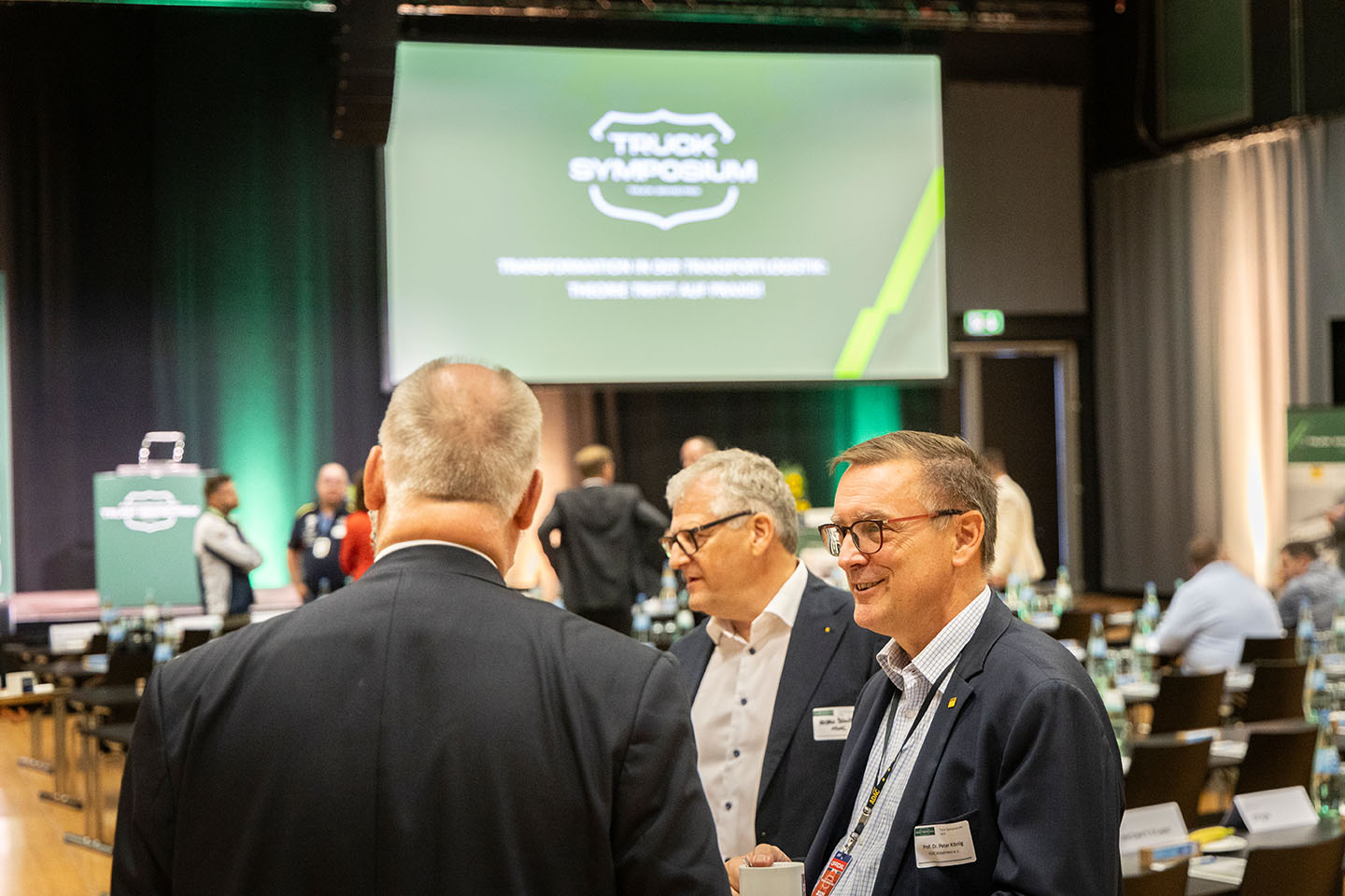 Eine Gruppe von Männern in Anzügen unterhält sich in einem Konferenzraum. Im Hintergrund ist eine große Projektionsfläche mit dem Logo „Truck Symposium“ und dem Thema der Veranstaltung zu sehen.