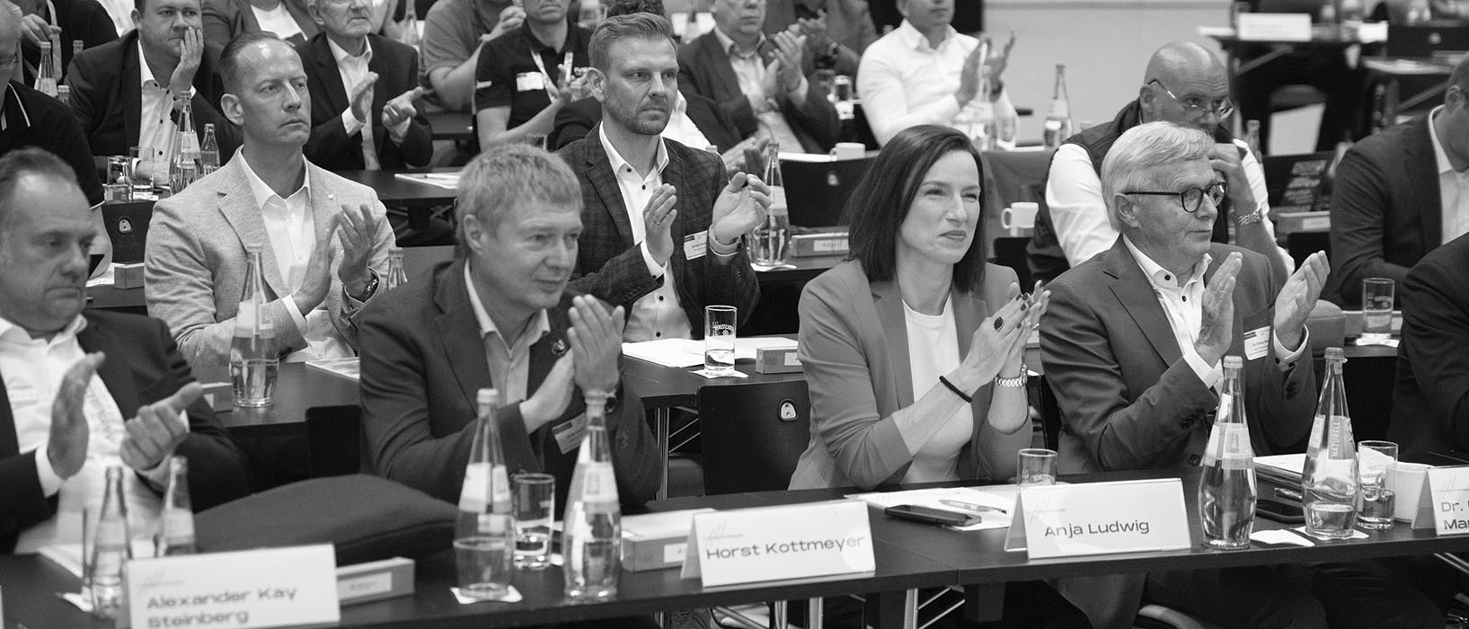 Eine Gruppe von Menschen sitzt an Konferenztischen und klatscht. Vor ihnen stehen Namensschilder und Getränkeflaschen. Die Stimmung wirkt konzentriert und respektvoll. Der Raum ist dicht besetzt mit mehreren Reihen von Zuhörern.