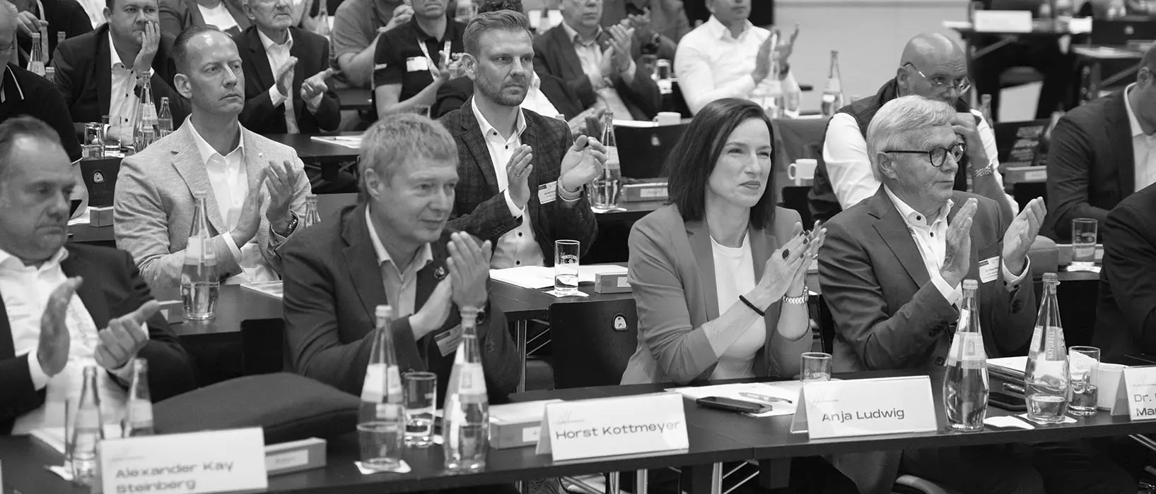 Eine Gruppe von Menschen sitzt an Konferenztischen und klatscht. Vor ihnen stehen Namensschilder und Getränkeflaschen. Die Stimmung wirkt konzentriert und respektvoll. Der Raum ist dicht besetzt mit mehreren Reihen von Zuhörern.